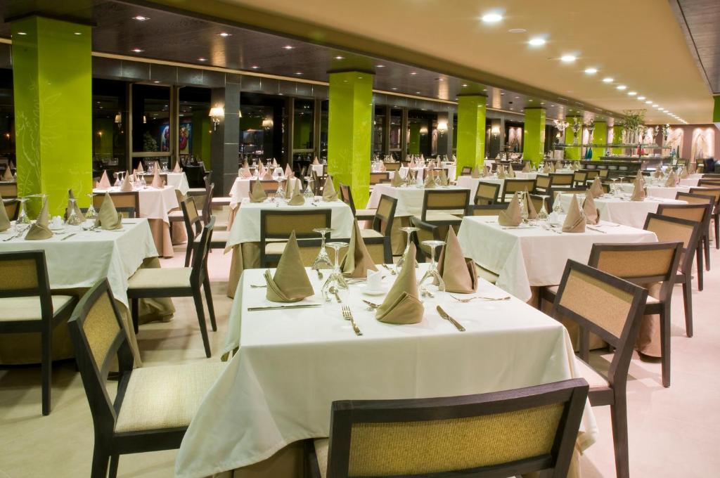 Labi iekārtots restorāns ar elegantām galdiem, baltiem galdautiem un izkārtotām galda piederumiem. Interjerā dominē zaļas kolonnas un silta apgaismojuma atmosfēra.