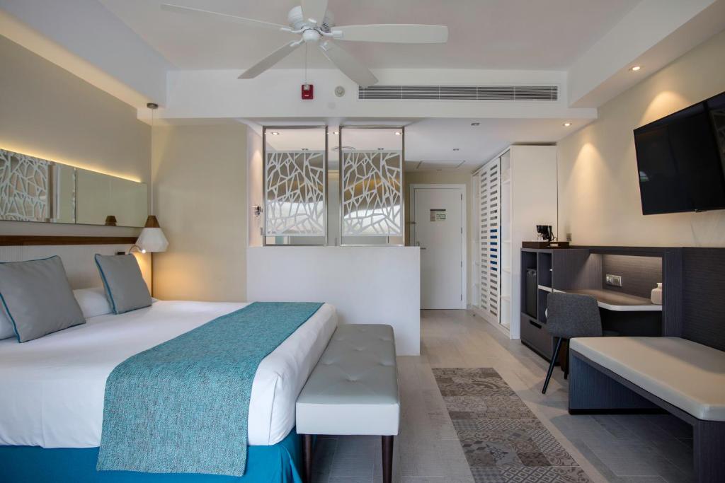 Mūsdienīga viesnīcas numura iekārtojums ar gultu, dekoratīvu segu un komfortablu atpūtas zonu. Telpa ir gaiša un eleganta, ar televizoru un atpūtas stūri.