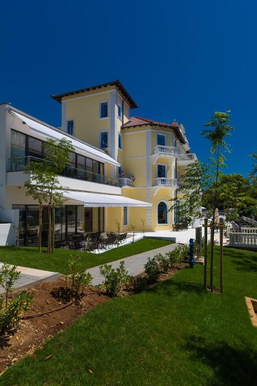 Mūsdienīga viesnīca ar dārzu un terasi, kurā redzamas zaļās zāles un jauni koki, zem skaidras zila debesīm.