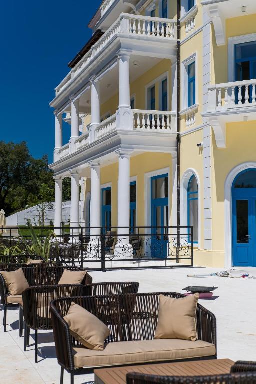 Elegantā viesnīcas ēka ar gaišu dzeltenu fasādi un zilu logu rāmjiem. Terasē redzamas modernas rotangpalmas mēbeles ar spilveniem, kas aicina atpūsties saulainā dienā.