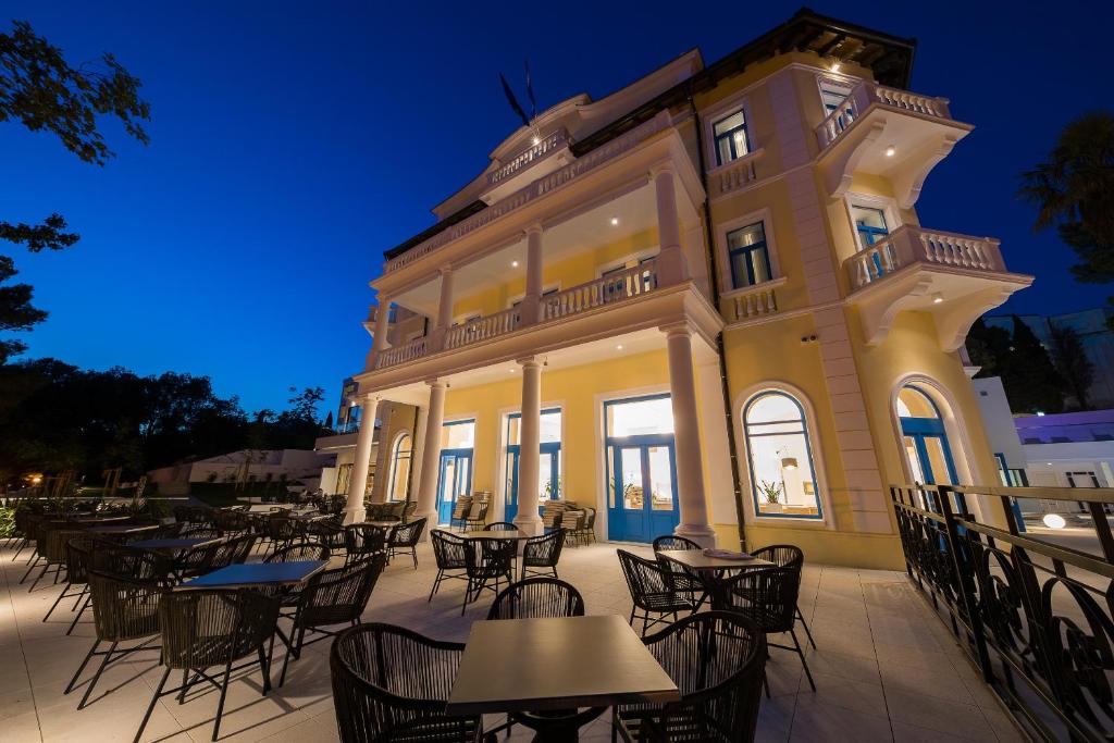 Elegantā ēka ar baltiem kolonām un zilu durvju krāsu, ap kuras izvietoti galdi un krēsli, radot patīkamu atmosfēru vakara pasēdēšanai. Gaismas izgaismota, ēka izceļas tumsā, piedāvājot viesiem patīkamu atmosfēru.