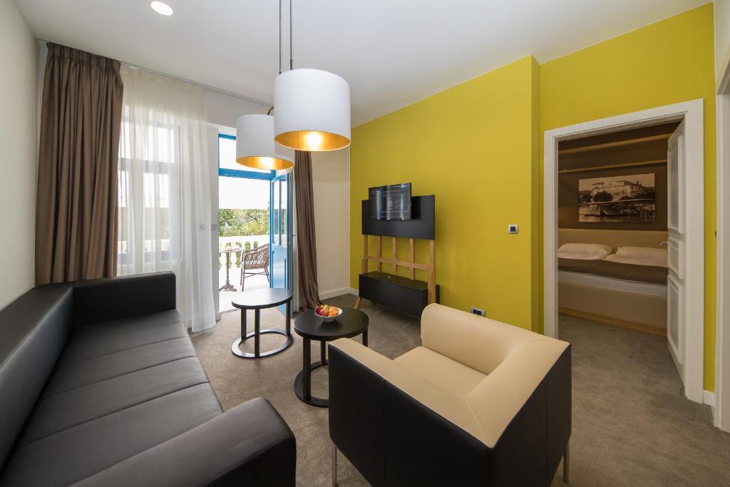 Mūsdienīgs viesnīcas numurs ar gaišām sienām, dīvānu un ērtu sēdvietu, kā arī balkona durvīm, kas ved uz āru. Interjerā ir televizors un minimālistiska dekorācija.