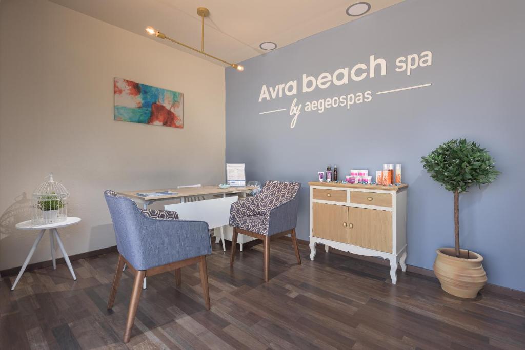 Mūsdienīgs spa interjers ar elegantiem krēsliem, galdu un dekoratīviem elementiem, kas rada relaksējošu atmosfēru. Uz sienas redzams spa nosaukums "Avra beach spa" ar stilīgu dizainu.