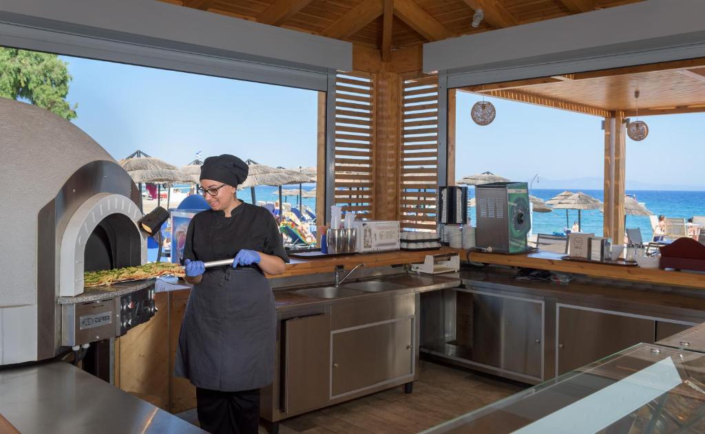 Pavārs, kas strādā pie picu cepeša krāsns pludmales restorānā, ar skatu uz jūru un saulainām pludmales gultām.