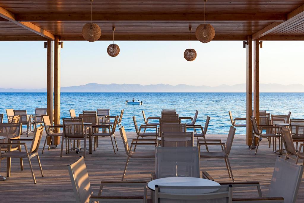 Jūras piekrastes restorāns ar koka konstrukciju un skatu uz ūdeni, kur redzami galdi un krēsli. Fons ar kalnu siluetu un zvejnieku laivu.