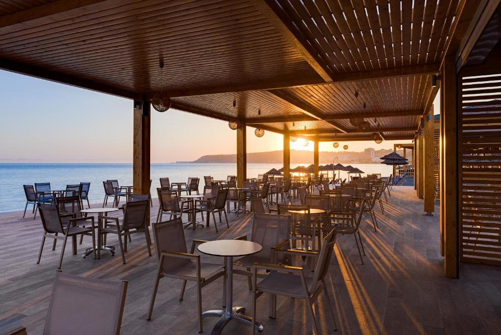 Jūras krastā esoša terase ar galdiem un krēsliem, kas apgaismota saulrieta gaismā. Skats uz mierīgu jūru un saules atspīdēšanu ūdenī, radot relaksējošu atmosfēru.