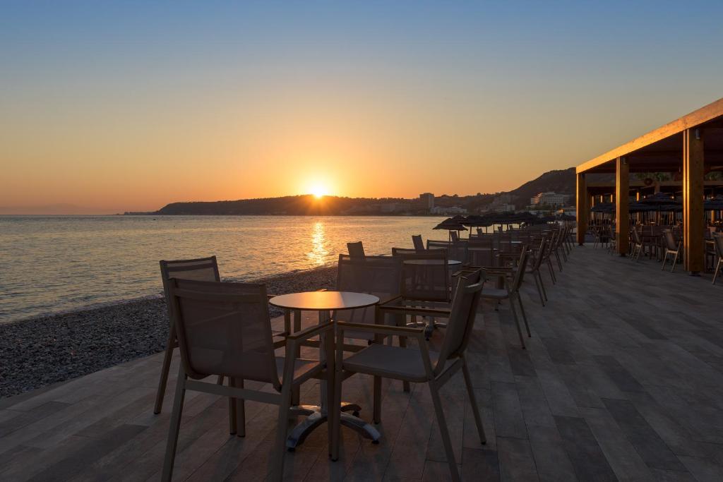 Saulriets pie pludmales kafejnīcas, kur redzami tukši galdi un krēsli, kas gaida apmeklētājus. Ūdens virsma atspoguļo saules gaismu, radot mierīgu atmosfēru.