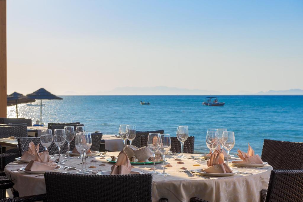 Eleganta restorāna galda kompozīcija ar glāzēm un galdautu, kas izvietots pie jūras ar skatu uz ūdeni un laivām. Ideāla vieta romantiskām vakariņām vai svinībām pludmalē.