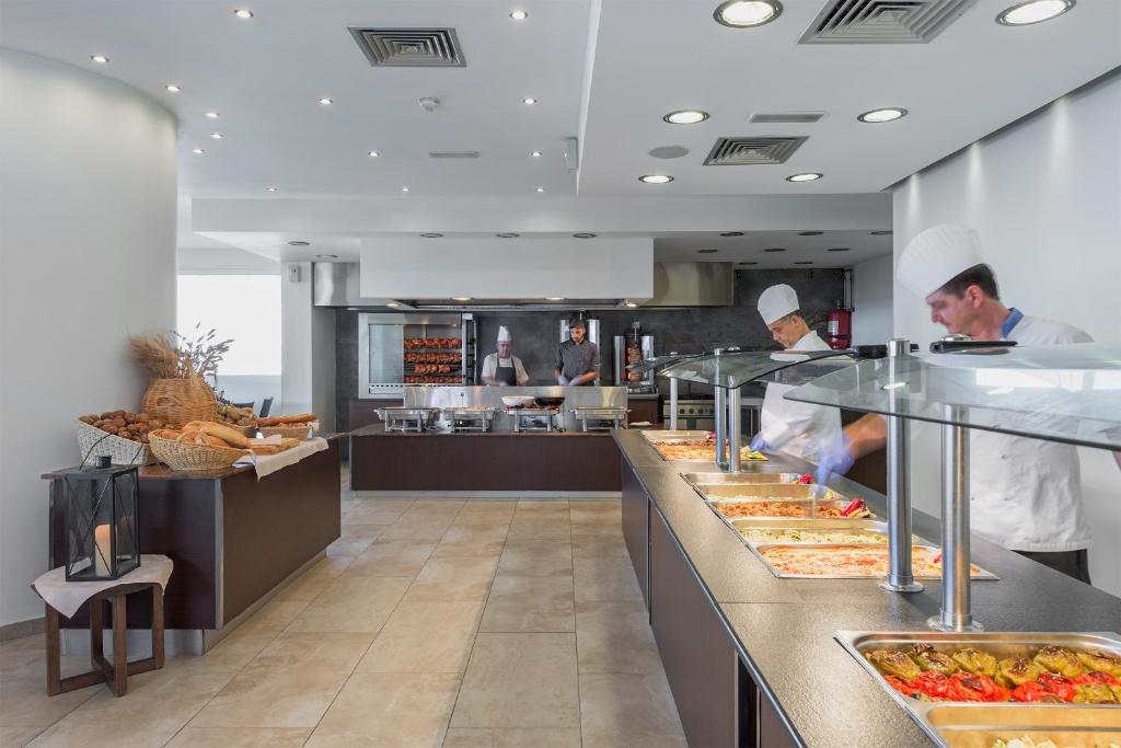 Mūsdienīga restorāna virtuve ar šefpavāriem, kas pasniedz dažādus ēdienus pie bufetes galda. Uz galda redzami vairāki ēdienu trauki un maizes grozs.