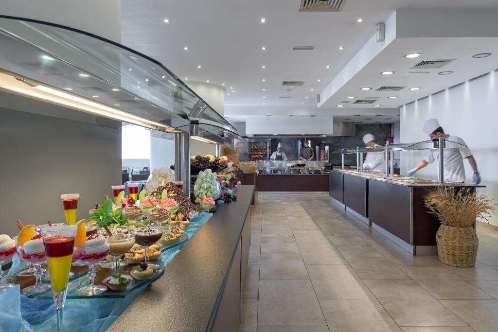 Deserts un konditorejas izstrādājumi izkārtoti uz galda, ar pavāriem, kas strādā virtuvē modernā restorānā. Gaismas un tīras līnijas veido patīkamu atmosfēru.