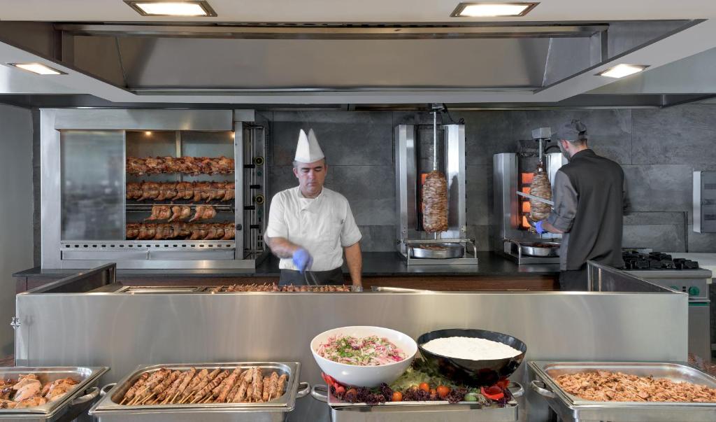 Virtuves skats ar pavāriem, kas gatavo gaļu uz grila, un dažādiem ēdieniem uz galda. Šajā modernajā restorānā redzami lieli gaļas gabali un svaigas salātu porcijas.