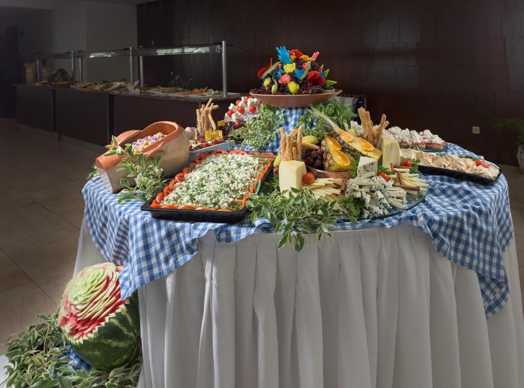 Svētku galda klājums ar dažādiem ēdieniem, ieskaitot sieru, augļus un salātus, rotāts ar zaļumiem un košiem elementiem. Galda virsma ir klāta ar zilu pārklāju un baltiem galdautiem.