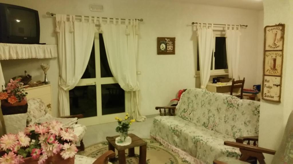 Mājīga viesistaba ar ziedu dekorācijām, mīkstiem dīvāniem un vintage televizoru, kas rada siltu un aicinošu atmosfēru. Logi ar vieglām aizkariem ļauj ieplūst dabiskajai gaismai.