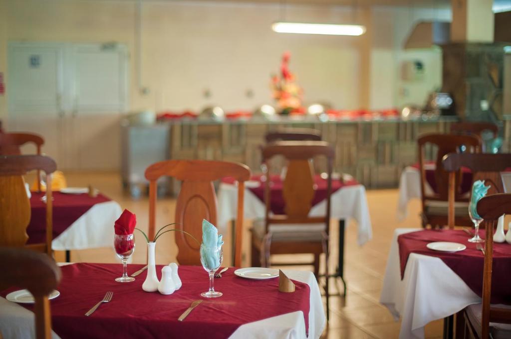 Restorāna interjers ar eleganti klātiem galdiem, sarkaniem galdautiem un dekoratīvām glāzēm. Fons ietver bufeti ar ēdieniem un ziedu kompozīciju.