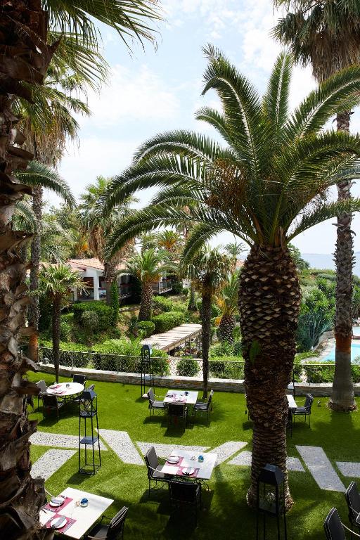 Tropiskā restorāna terase ar palmām un galdiem, kas izvietoti zaļā zālienā. Skats uz dārzu un baseinu, radot atpūtas atmosfēru.