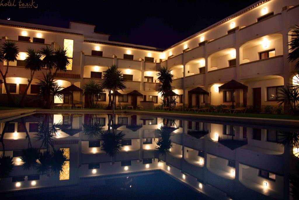 Nakts skats uz viesnīcas ēku ar apgaismojumu un zilu baseinu, kas atspoguļo apkārtējo ainavu.