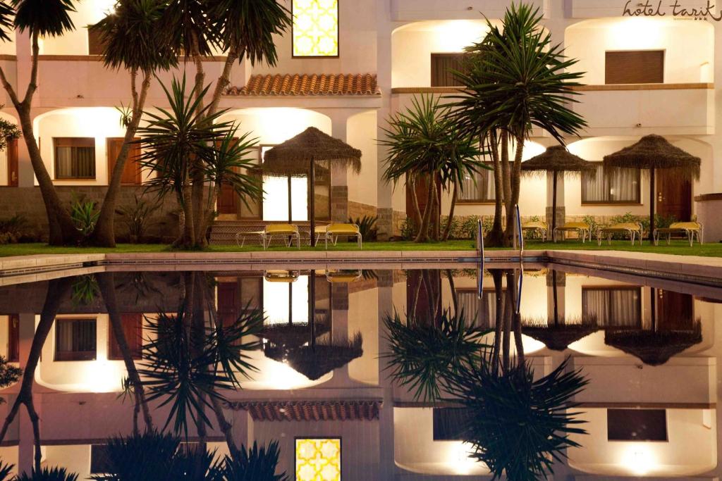 Nakts ainava ar baseinu, kurā atspoguļojas viesnīcas ēka ar palmām un sauļošanās krēsliem, radot mierīgu un relaksējošu atmosfēru.