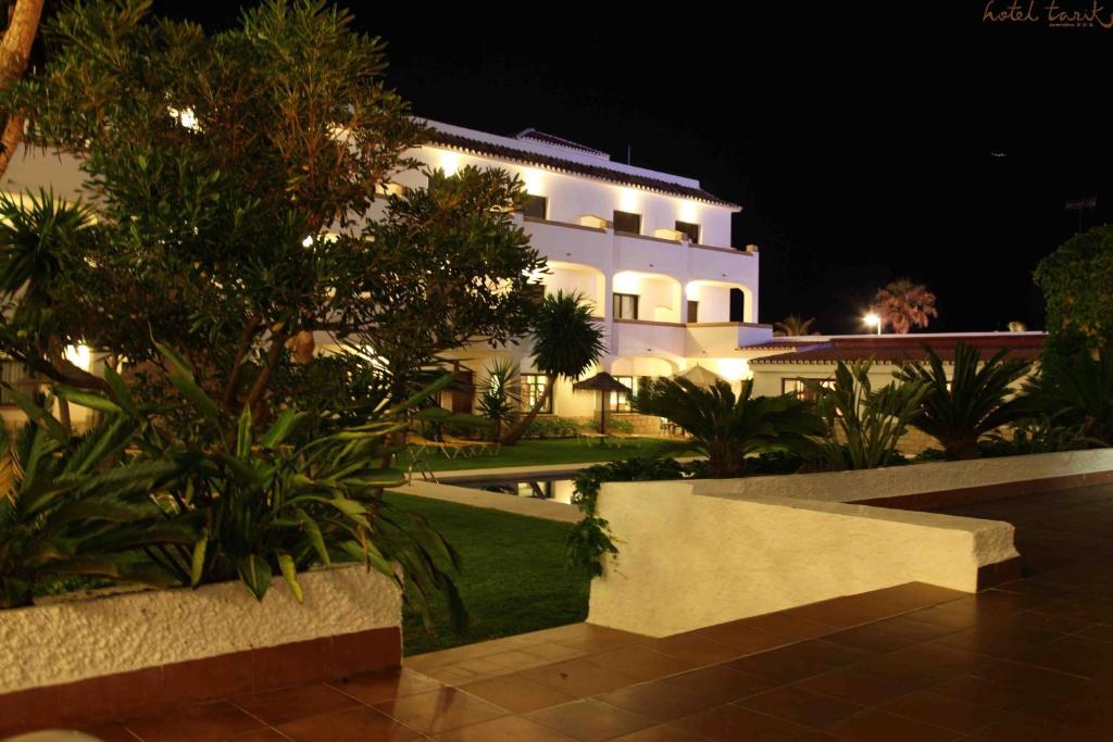 Nakts ainava ar viesnīcu, kas apgaismota ar mīkstu gaismu, apkārt esošās zaļās augu kompozīcijas un mierīga dārza atmosfēra.