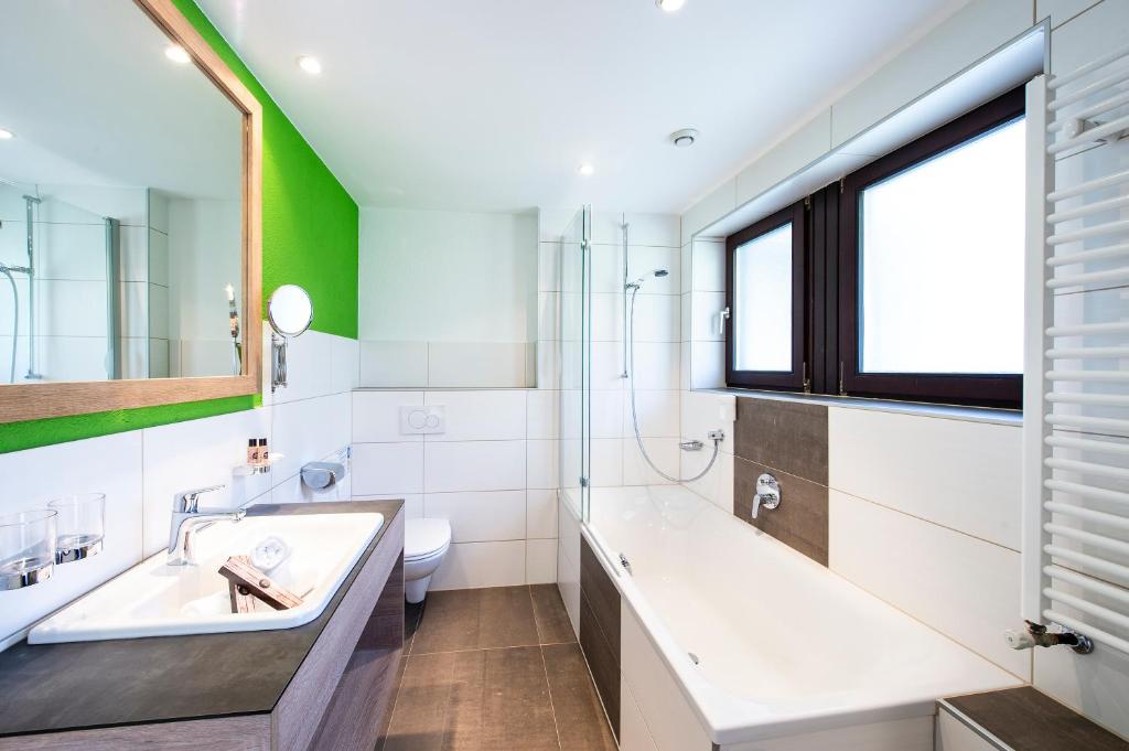 Mūsdienīga vannas istaba ar vannu, dušu un gaišām flīzēm, izceļot zaļas sienas akcentu un elegantu spoguli. Ideāla telpa relaksācijai un komfortam.
