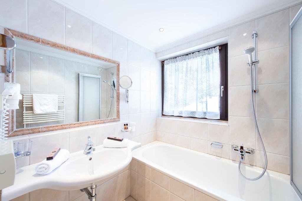 Mūsdienīga vannas istaba ar gaišām flīzēm, vannu un izsmalcinātu spoguli. Telpā ir arī dušas kabīne un logu ar baltu aizkaru.