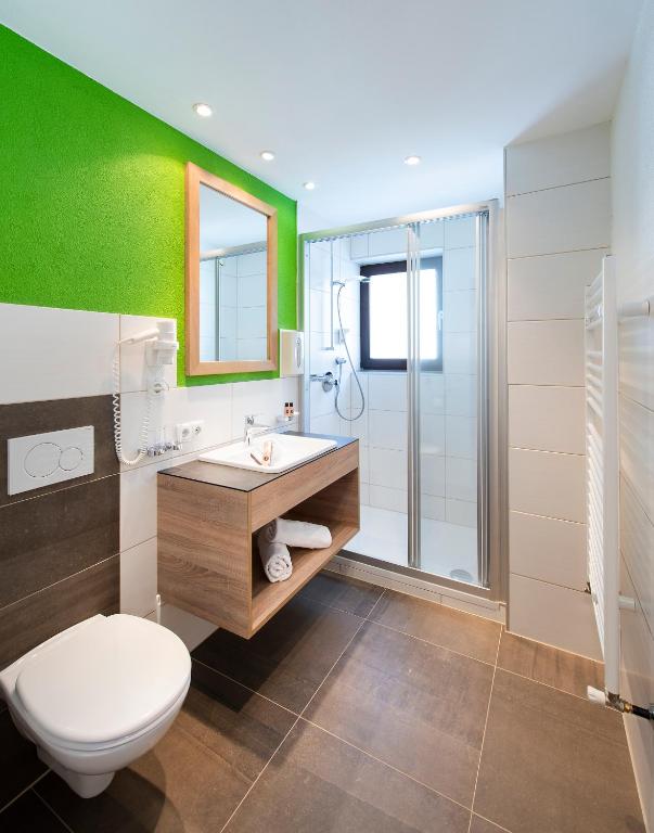 Mūsdienīgs vannas istabas interjers ar zaļu sienu, koka izlietni un stikla dušas kabīni. Vannas istaba ir gaiša un funkcionāla, nodrošinot komfortu un stilu.