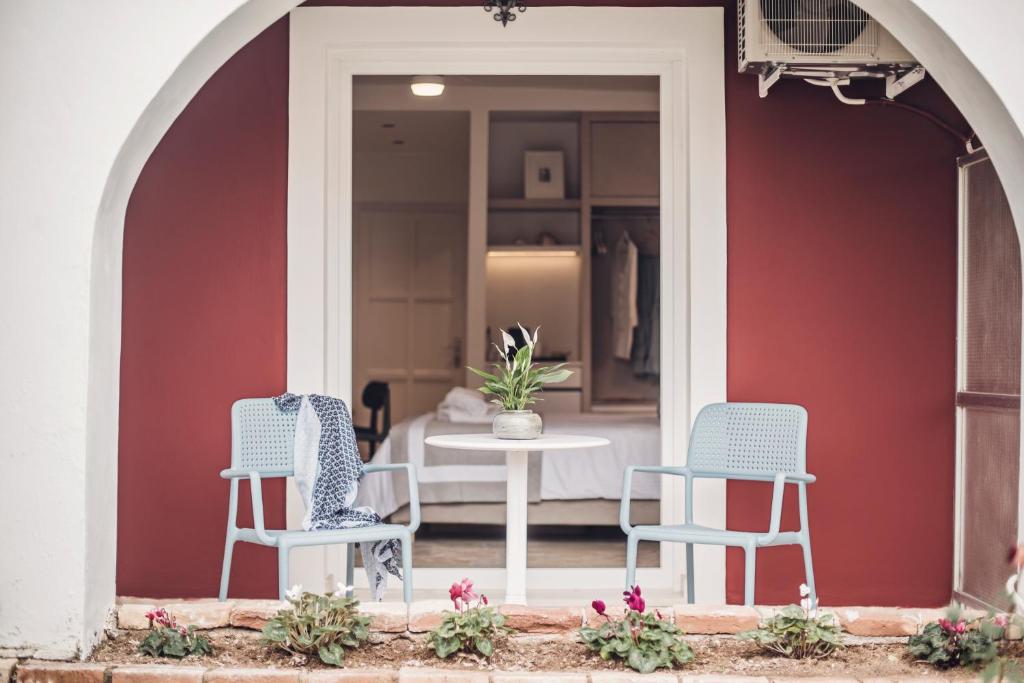 Mierīga terase ar divām gaisīgām krēsliem un mazu galdiņu, kas izvietota pie guļamistabas durvīm. Fons ir siltā sarkanā krāsā, un terasi rotā ziedi.