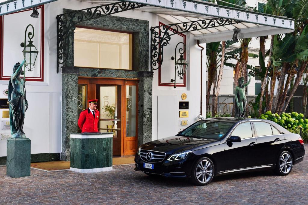 Greznas viesnīcas ieeja ar melnu Mercedes automašīnu un viesmīli sarkanā uniformā. Apkārt ir zaļi augi un skulptūras, radot elegantu atmosfēru.