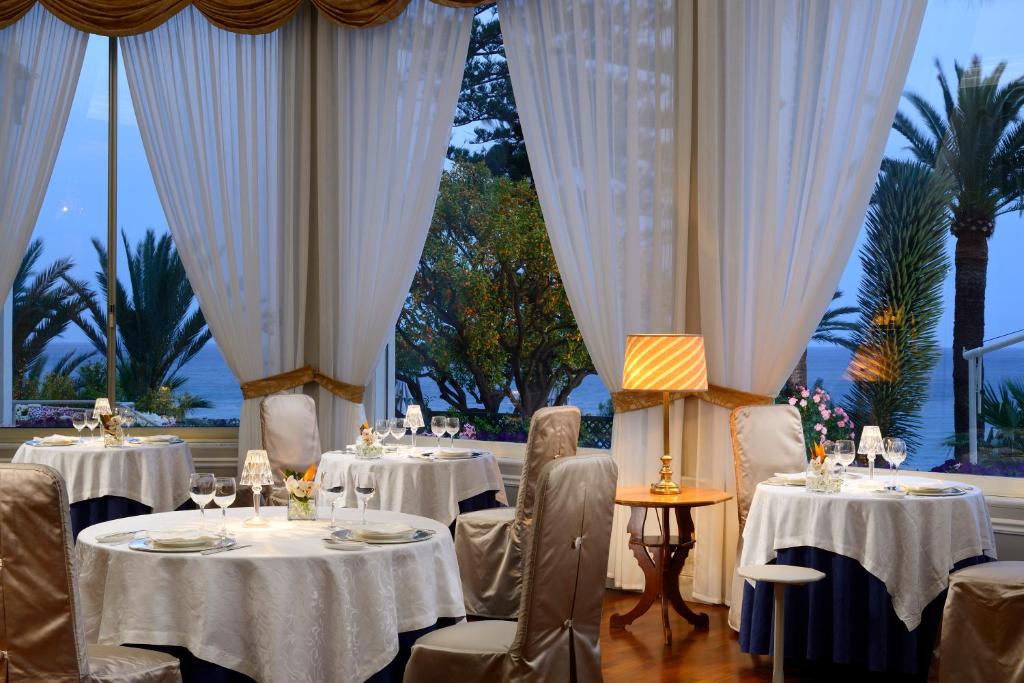 Elegants restorāns ar skatu uz jūru, kur baltas galdautas un izsmalcināti galda piederumi veido patīkamu atmosfēru. Lielie logi ļauj ienākt dabiskai gaismai un piedāvā skatu uz palmu kokiem ārpusē.