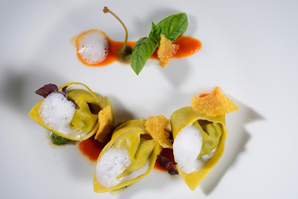 Mūsdienīgs ēdiens ar krāsainām ravioli, rotāts ar putām un garnējumu, pasniegts uz balta šķīvja.