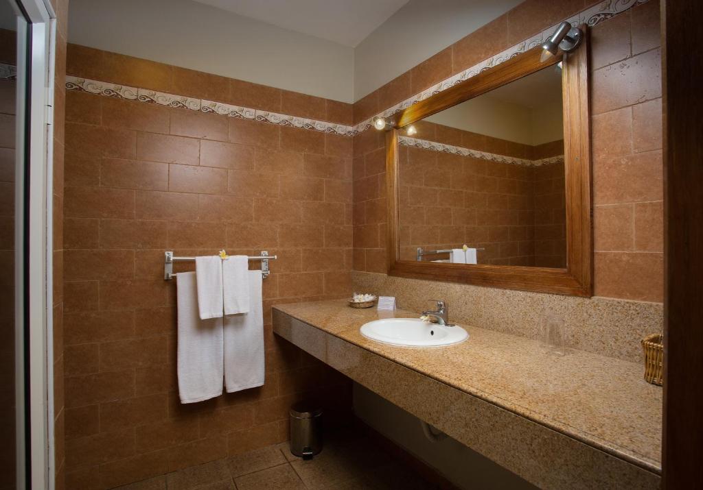 Mūsdienīga vannas istaba ar brūnu flīžu apdari, plašu izlietni un spoguli, kas nodrošina gaismu. Divas baltas dvieļi un aksesuāri uz letes.