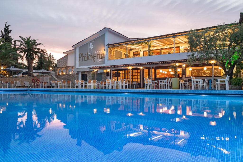 Viesnīcas "Philoxenia" ēka ar baseinu un terasi, kas apņemta ar palmām un zaļumiem, radot relaksējošu atmosfēru. Vakara apgaismojums piešķir īpašu burvību.