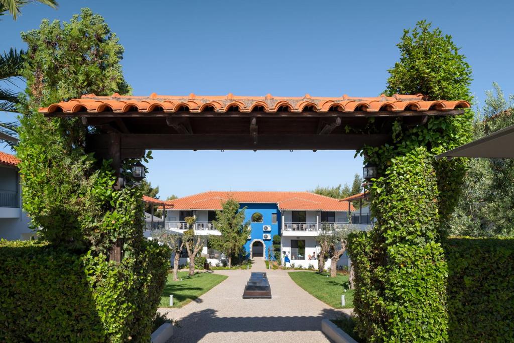 Skaista ainava ar ieeju, kas ietver zaļus krūmus un akmens celiņu, kas ved uz ēku ar sarkanu jumtu un zilām sienām. Dārzs ir labi iekopts un aicinošs.