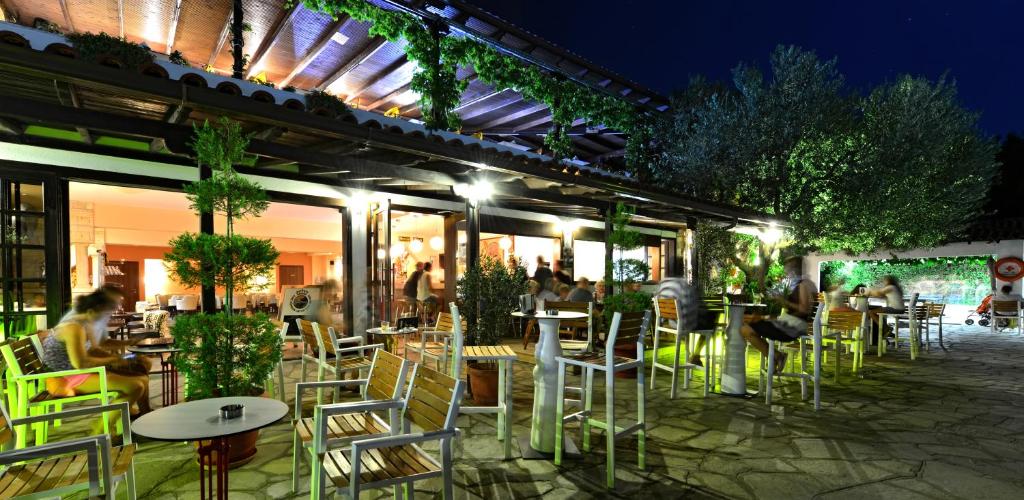 Āra kafejnīca ar zaļu apstādījumu un apgaismojumu, kas rada patīkamu atmosfēru vakara stundās. Apmeklētāji relaksējas uz terases, baudot dzērienus un ēdienus.