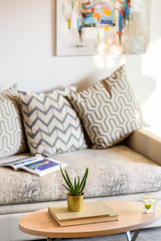 Mūsdienīgs dzīvojamās istabas stils ar dīvānu, dekoratīvajām spilveniem un mazu augu uz galda. Gaisma ieplūst telpā, radot mājīgu atmosfēru.