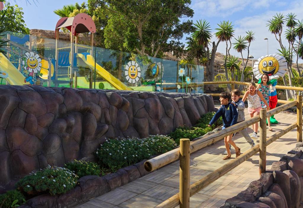 Bērni izbauda jautru brīdi rotaļu laukumā ar slidkalniņiem un krāsainām figūrām. Apkārt redzamas zaļas augu kompozīcijas un saulaini dekori.