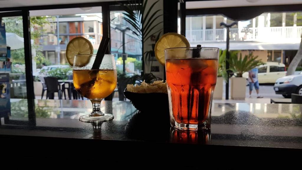 Divi dzērieni, viens ar citrona šķēli un zaļo stiebru, otrs dzidrs un sarkans, novietoti uz galda ar skatu uz āra terasi.