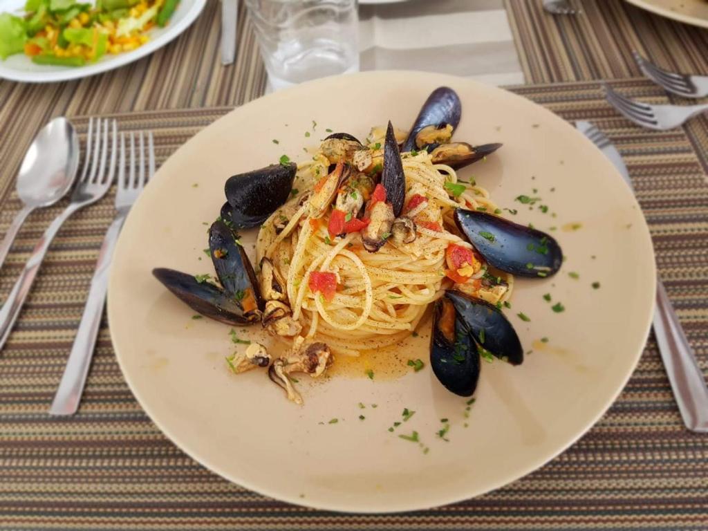 Pasta ar mīdijām, garnelēm un tomātiem, rotāta ar zaļumiem, pasniegta uz krēmkrāsas šķīvja.