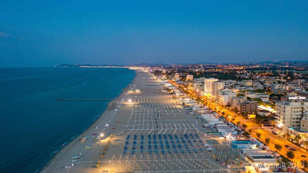 Naktī uzņemta ainava, kurā redzams pludmales krasts ar saulessargiem un gultām, gar kuru stiepjas pilsētas iela ar apgaismojumu. Jūras viļņi un nakts debesis veido mierīgu atmosfēru.