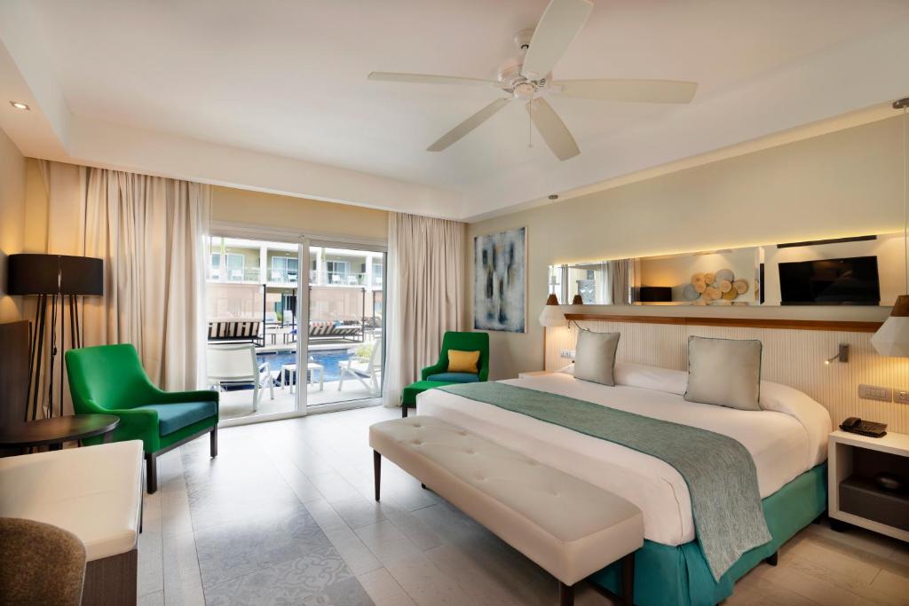 Mūsdienīga viesnīcas numura interjers ar plašu gultu, krāsainām sezonas krēslām un piekļuvi baseinam. Gaisma plūst caur lielajiem logiem, radot patīkamu atmosfēru.