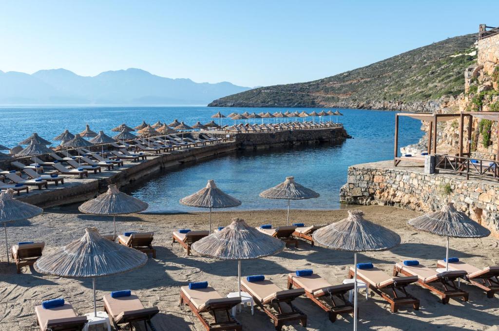 Aquila Elounda Village pludmales skats ar sauļošanās krēsliem un lietussargiem piekrastē.