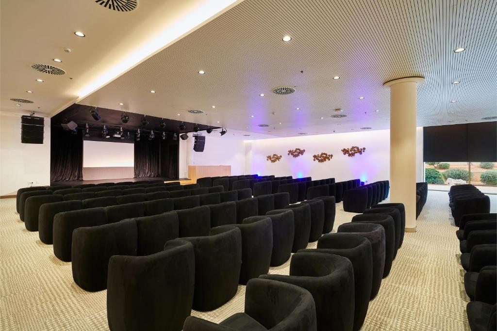 Konferenču zāle ar melniem krēsliem, skatuves apgaismojumu un ekrānu prezentācijām. Telpa ir moderna un piemērota pasākumiem.