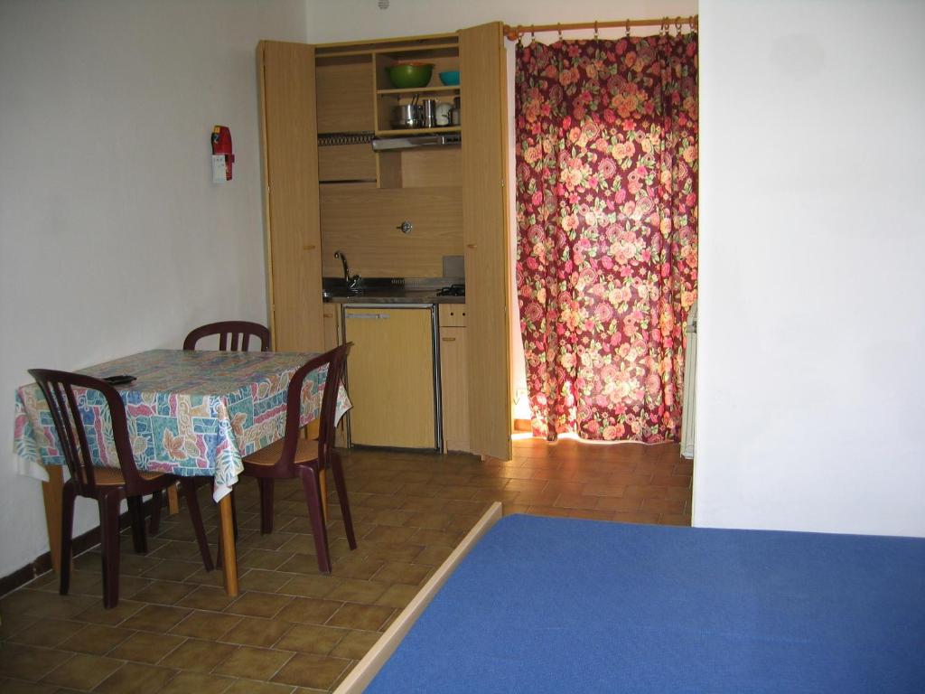 Kompakta virtuve ar galdu un krēslām, kas izvietota istabā. Aiz aizkaru ar ziedu rakstu redzama virtuve ar plauktiem un traukiem.