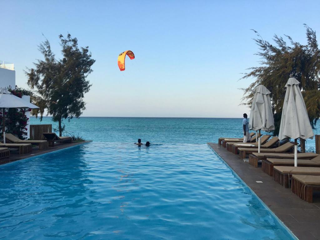 Skats uz baseinu pie pludmales, kur divi cilvēki peld, bet uz ūdens lido kitesurferis ar krāsainu burtu. Fons veido mierīgu jūras ainavu ar zilu debesi un zaļām palmām.