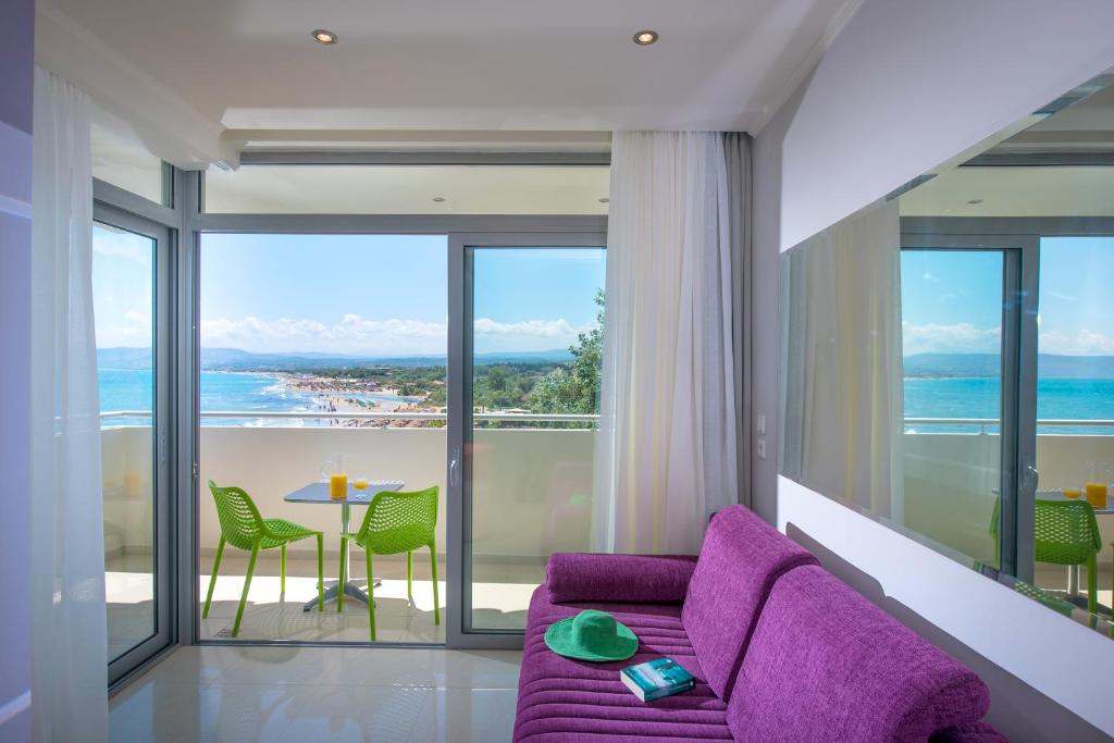 Mūsdienīgs interjers ar skatu uz jūru, kurā redzams rozā dīvāns un zaļas krēsli pie galda. Aiz logiem izskatās pludmale un zila jūra.