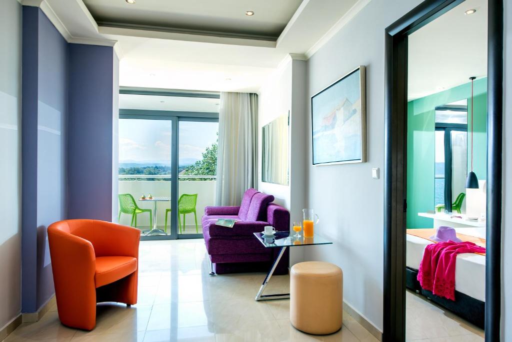Mūsdienīgs viesnīcas numurs ar spilgtiem mēbeļu akcentiem, ietverot purpura dīvānu un oranžu krēslu, kā arī skatu uz terasi ar zaļām krēsliem. Gaismas un krāsas rada patīkamu atmosfēru.