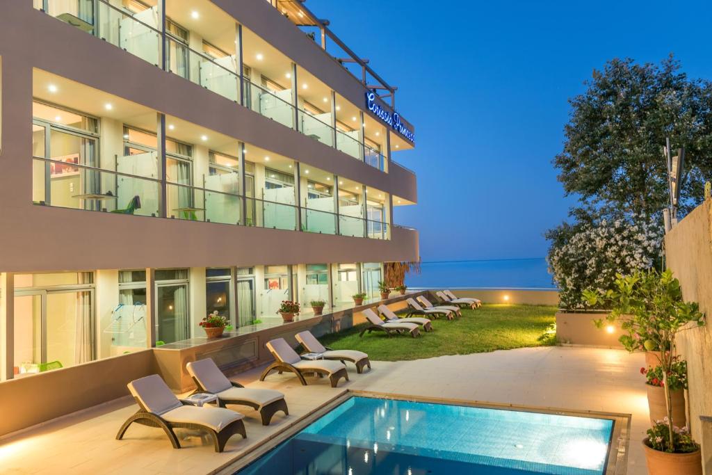 Mūsdienīgs viesnīcas komplekss ar baseinu un sauļošanās krēsliem, blakus jūrai un zaļai zālei. Vakara apgaismojums piešķir vietai idillisku atmosfēru.