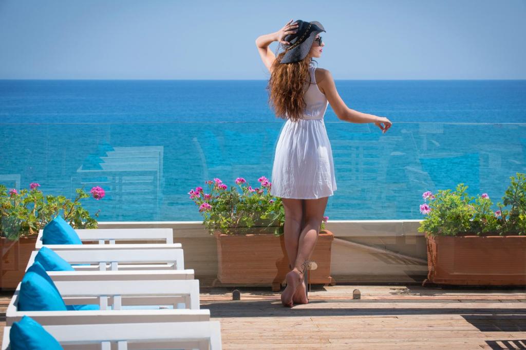 Sieviete baltā kleitā stāv uz terases ar skatu uz jūru, baudot sauli un dabu. Ap viņu ir ziedu podi un ērtas atpūtas krēsli.