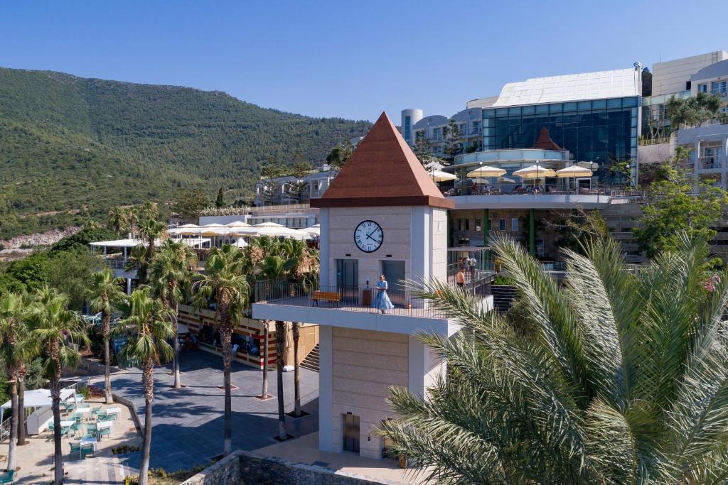Viesnīcas Duja Bodrum pulksteņa tornis piekrastē ar palmām un kalniem fonā.