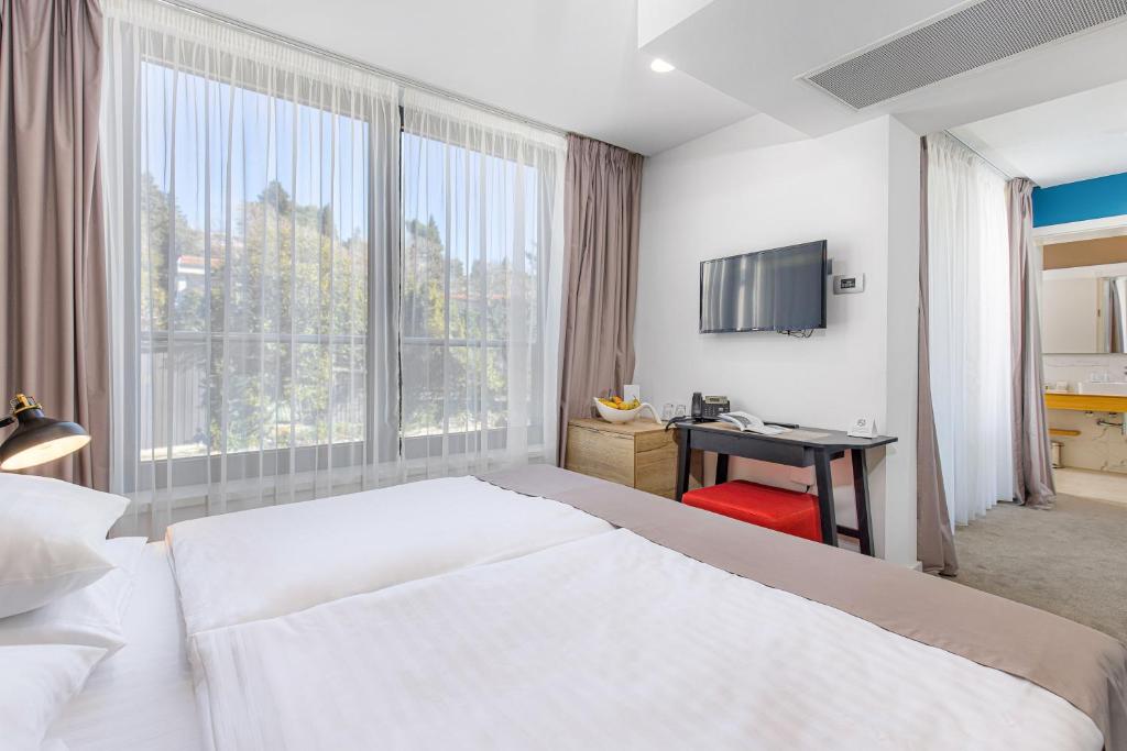 Mūsdienīga viesnīcas numura iekārtojums ar plašiem logiem, gultu un televizoru, kas nodrošina komfortablu atpūtu un gaišu atmosfēru.