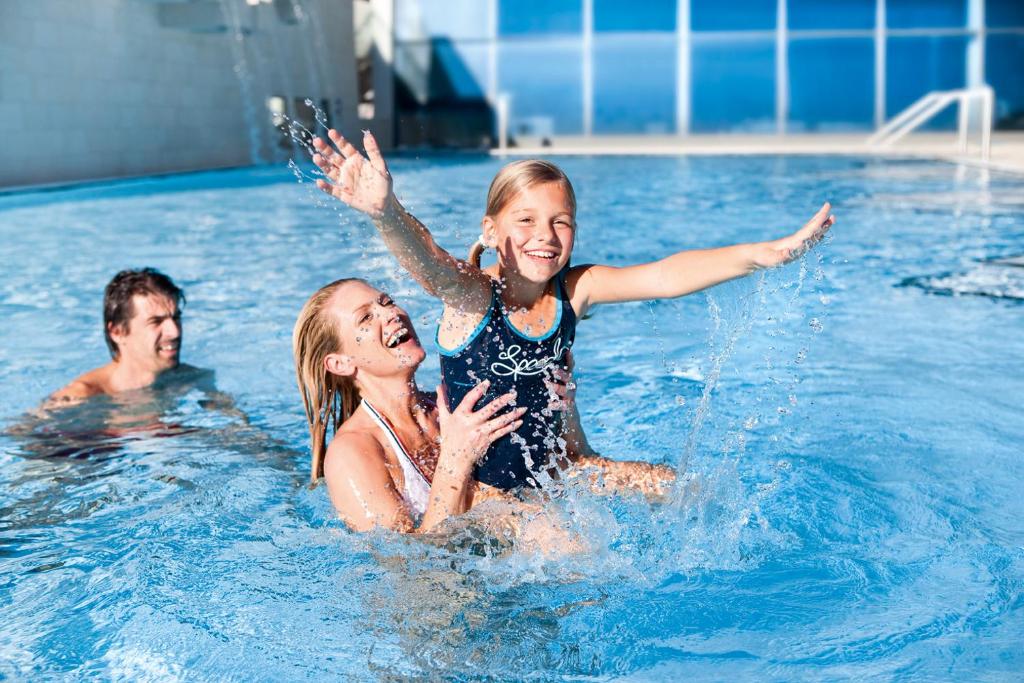 Ģimenes prieki peldbaseinā, kur māte un meita kopā izbauda ūdens spēles, kamēr tēvs peld tuvumā.